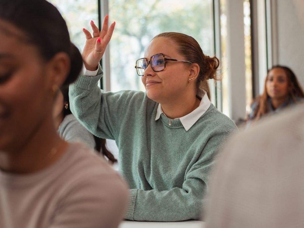 Etudiante de l'ESGCI blonde avec des lunette et un pull vert dans une salle de classe lève la main