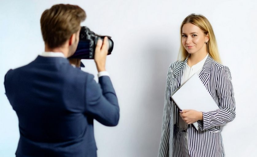 Jeune femme blonde en tenue professionnelle sur un fond blanc avec un homme photographe brun de dos avec un appareil photo